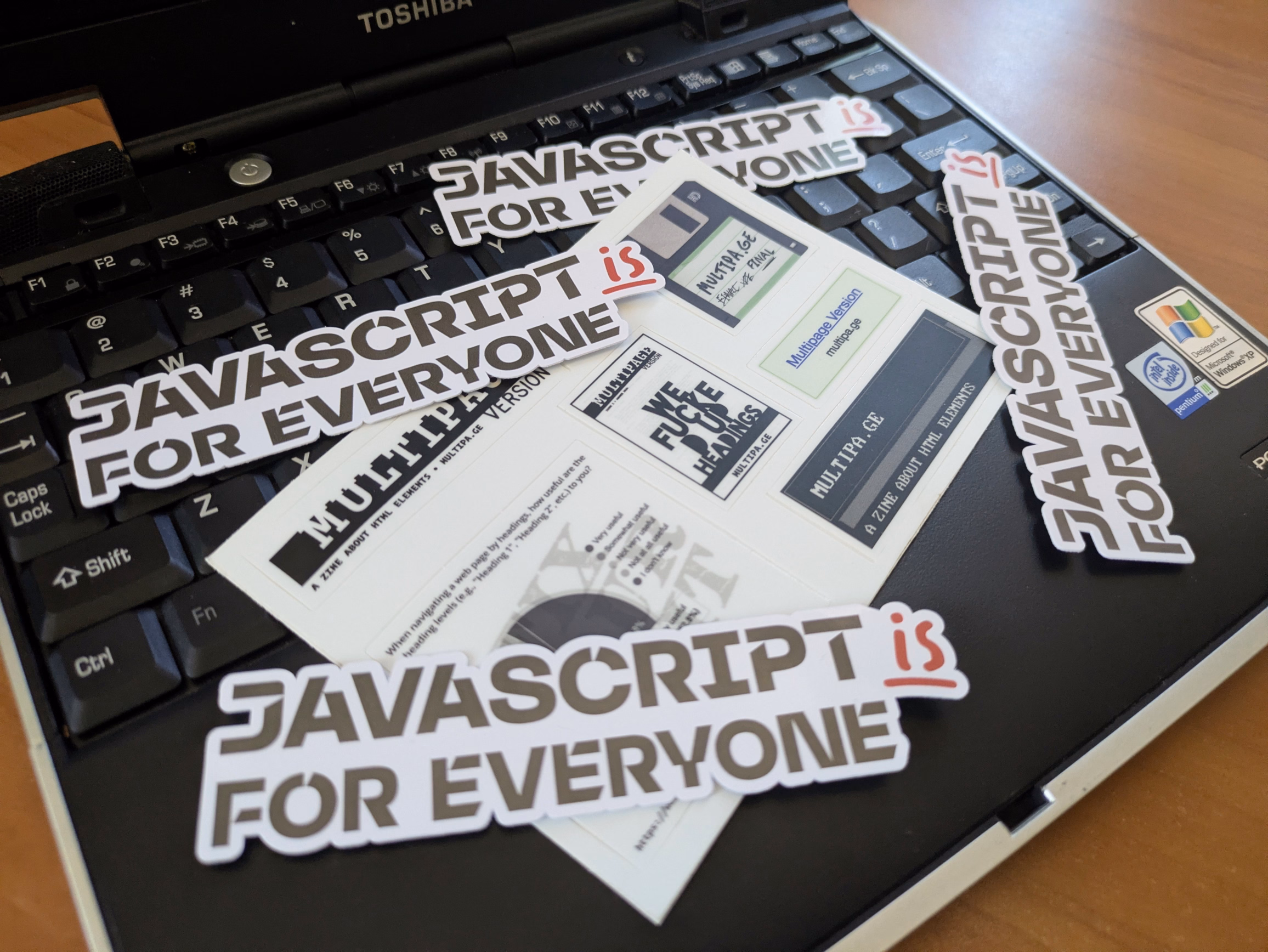 A collection of stickers reading 'JavaScript is for Everyone' and a sheet of Multipage Version stickers splayed on an old laptop keyboard.