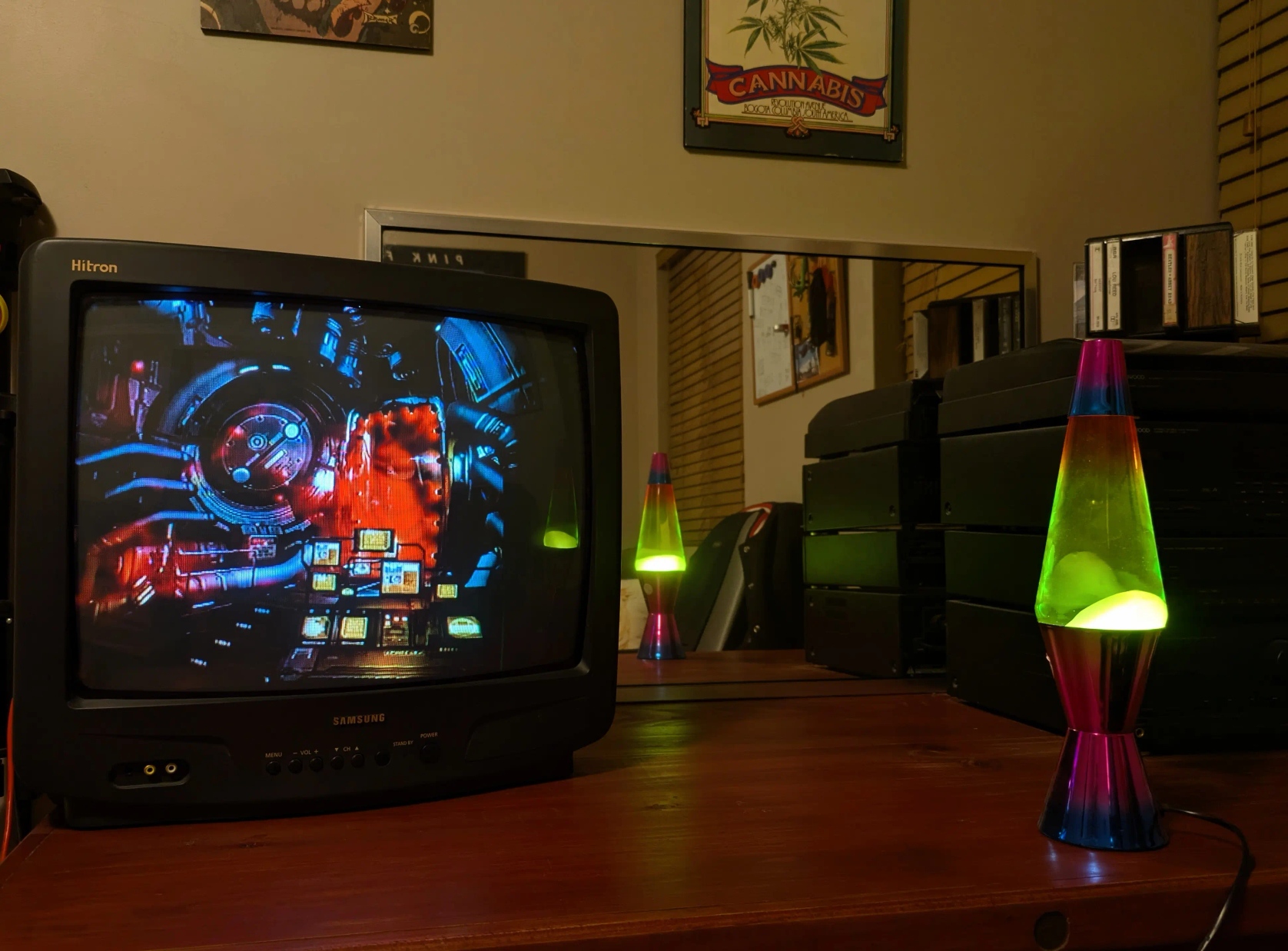 A 90s Samsung Hitron, a lava lamp, and a HiFi system on a wooden desk. A Doctor Strange poster and a cannabis poster can be seen in the background. Cassettes sit atop the HiFi and at the back of the desk is a mirror reflecting a Pink Floyd poster.