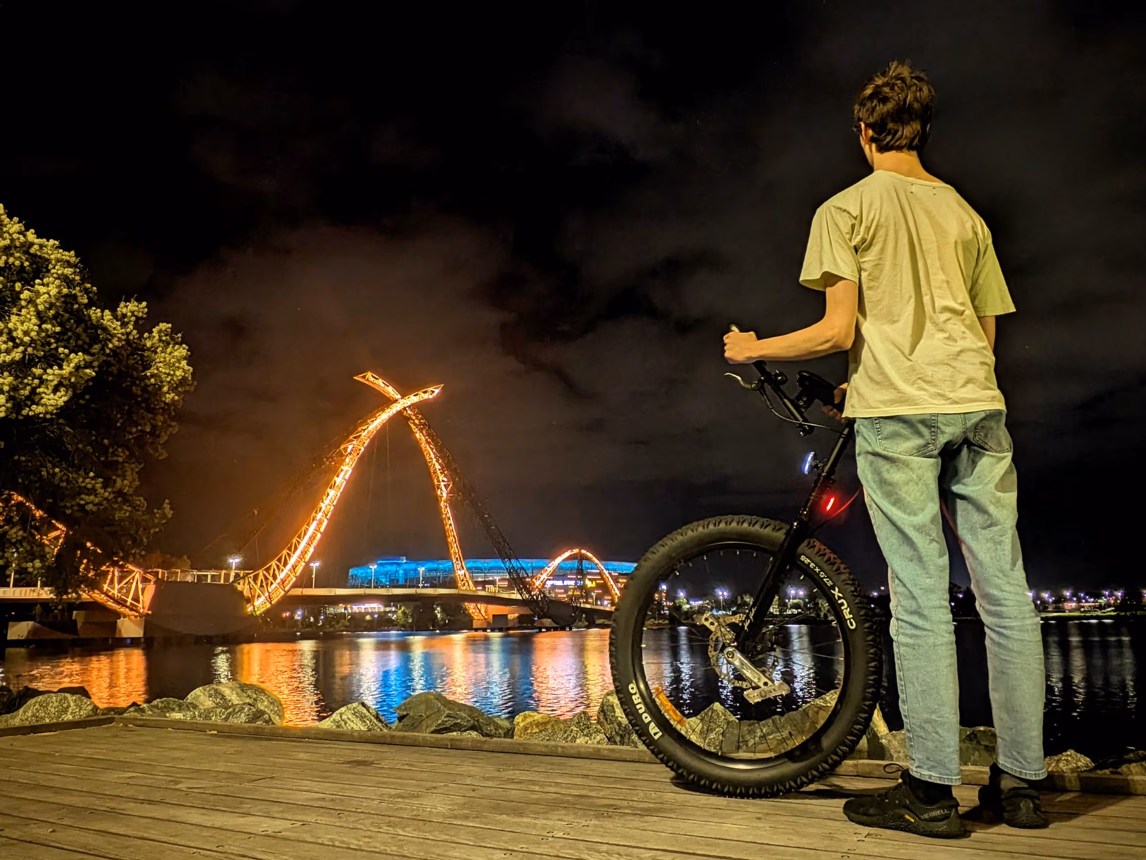 A bright, illuminated bridge that coils into the sky in front of a massive stadium at night. It is reflected in a river. In the foreground is me beside my unicycle.