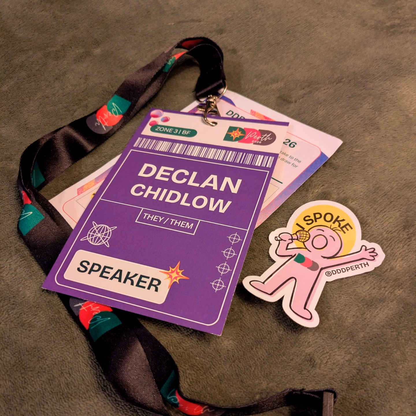 A lanyard with a card reading 'Declan Chidlow. Speaker.'. Beside it is a sticker with an illustration of a person with a microphone and the text 'I spoke @DDDPerth'.